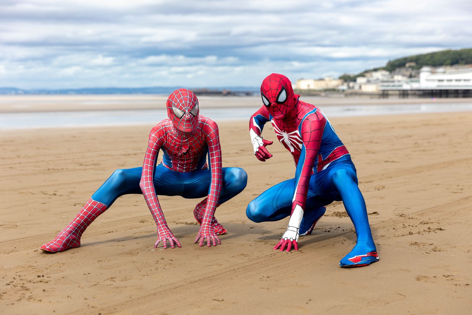 Two figures in Spidermen costumes in crouched down Spiderman positions on a beach